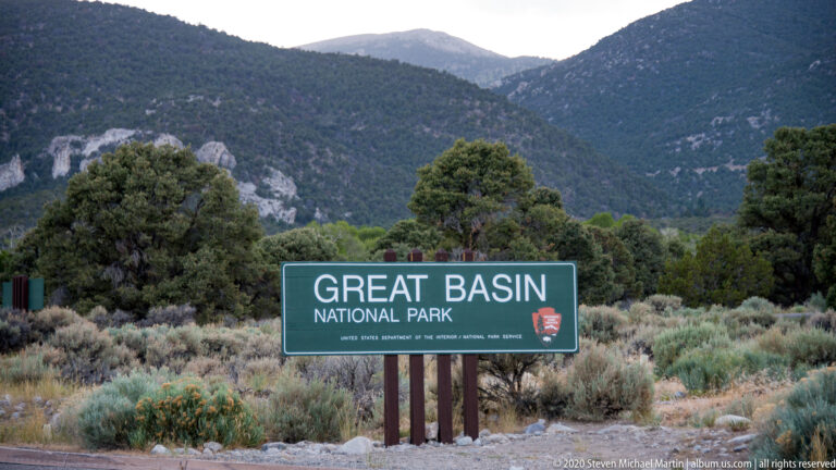 A green Great Basin National Park sign with an NPS emblem stands before wooded mountains.