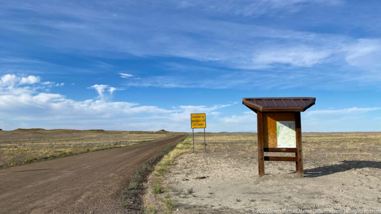 Dirt access road and information kiosk stand in open high-desert grassland beneath a broad blue sky.