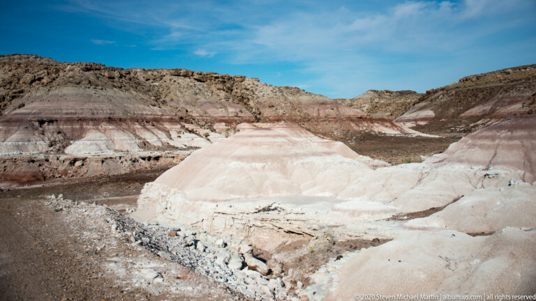 A pale, layered sediment mound fills the foreground beneath a dark eroded ridge and blue sky.
