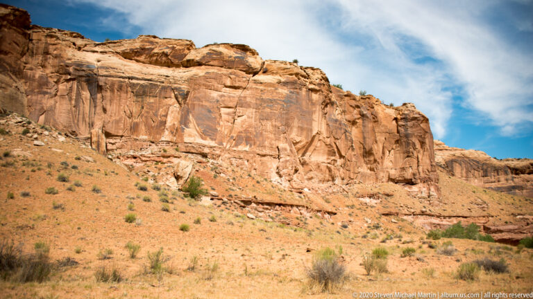 A tall sandstone wall stands above a sparse slope beneath streaked clouds.