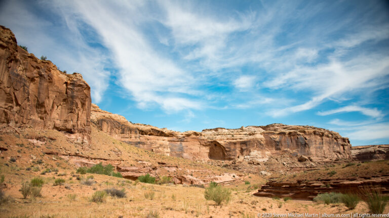 A sandstone wall borders an open wash dotted with shrubs beneath a cloud-streaked sky.