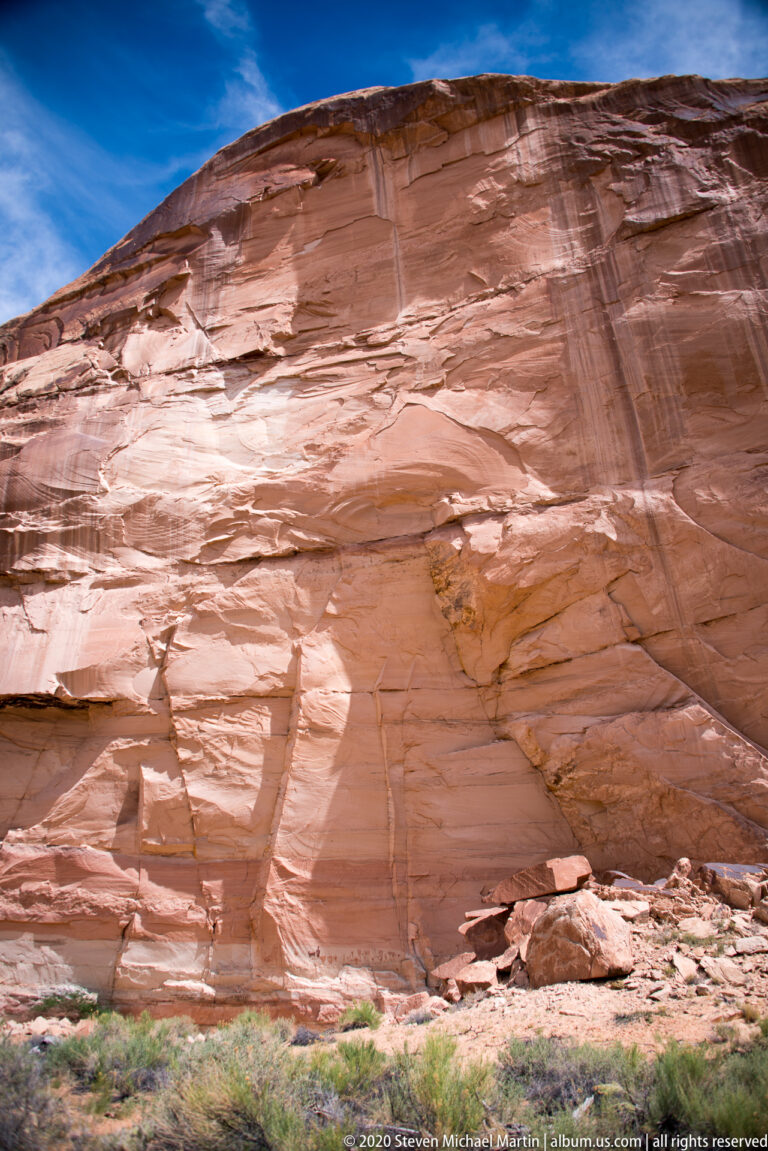 A smooth banded sandstone cliff with dark runoff streaks rises above a rocky base.