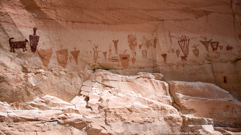 A long band of faded pictographs spans a sandstone shelter above a broad rock bench.