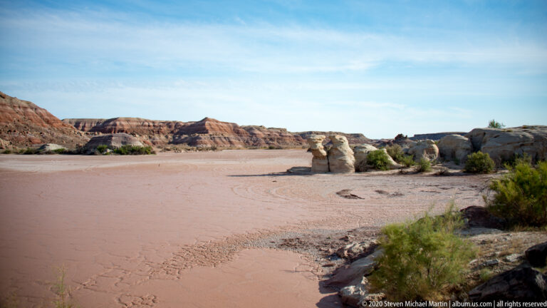 A solitary sandstone pillar stands beyond a pale dry wash, with layered red ridges across the background.