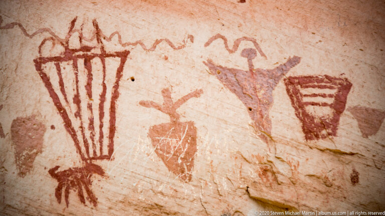 A pale sandstone wall bears red fan-shaped marks, a purple human-like figure, and a small barred motif.