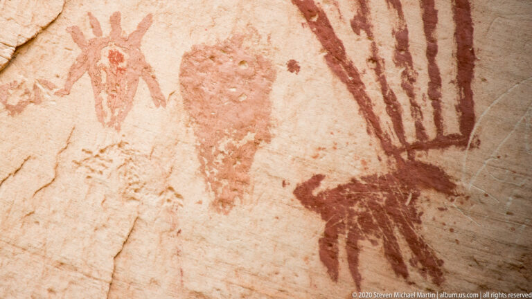 A dark red fan-shaped pictograph dominates a close sandstone view, with faint figures and lines at left.