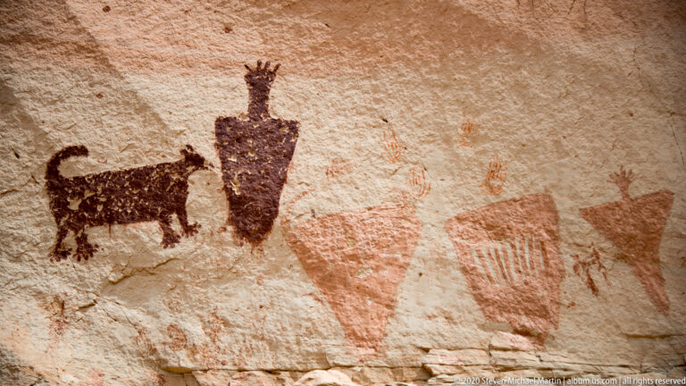 A dark four-legged animal and tall upright figure appear beside faded triangle and barred marks on sandstone.