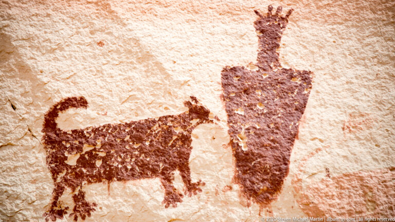 A dark animal figure with a curled tail stands beside a tall tapered figure on sandstone.