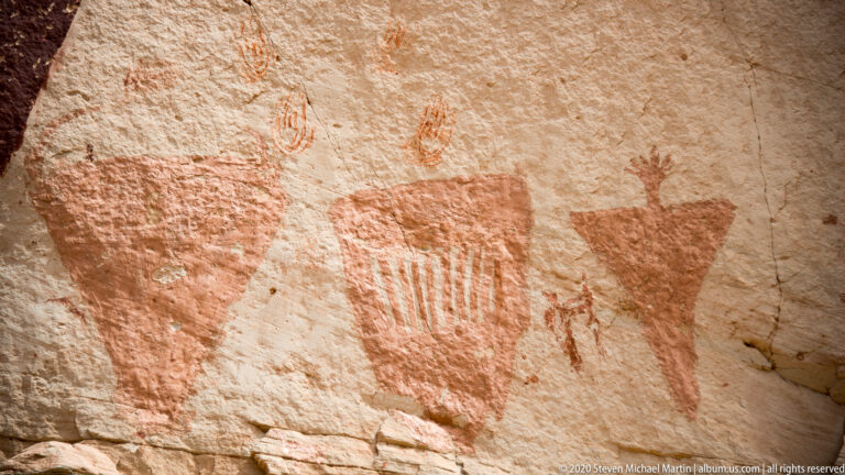 A broad triangle, barred central form, and smaller tapered motif span a pale sandstone wall.