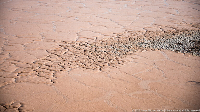 Polygonal cracks and rough, broken fragments pattern a dry pink-tan clay surface.