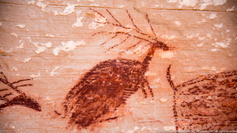 A dark tapered painted figure with branching lines sits between partial animal forms on sandstone.