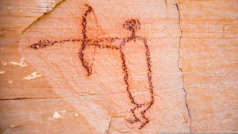 A thin outlined human-like figure extends both arms beside a vertical crack in sandstone.