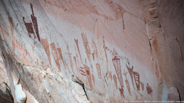 A person stands below a sloping sandstone wall lined with faded red pictographs.
