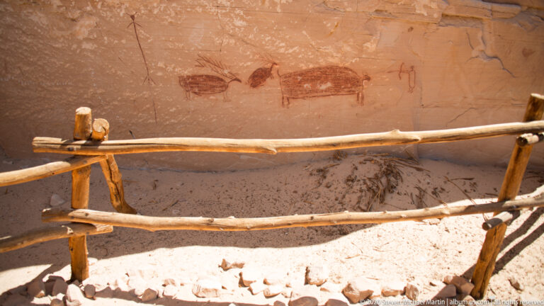 A lone hiker crosses a sandy clearing toward a massive orange sandstone alcove and pale cliff bands.