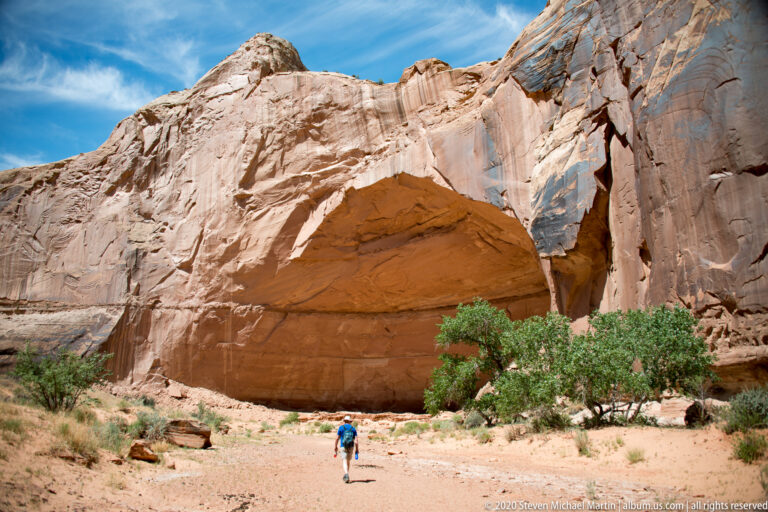 A sandy path winds between a dark shaded wall and a sunlit sandstone face toward an open canyon.