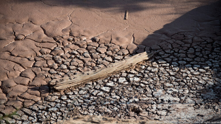 A weathered branch lies across broken clay plates amid broad polygonal cracks in a dry basin.