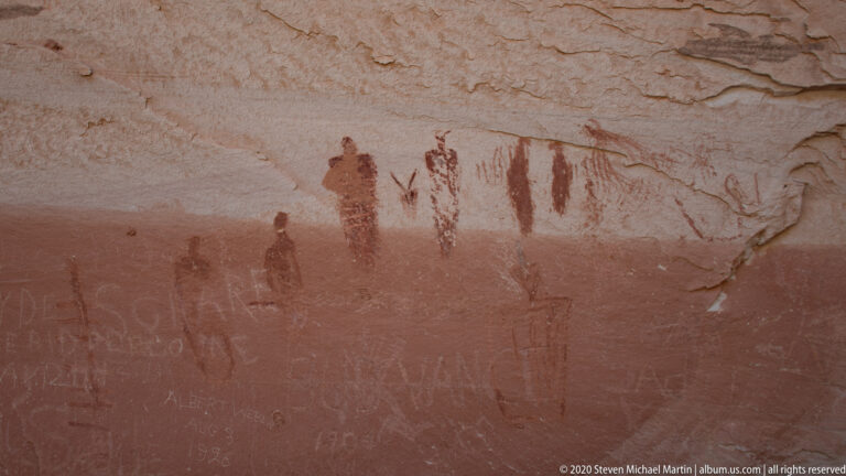 A dark painted figure and faint lines sit above pale scratched lettering on orange sandstone.