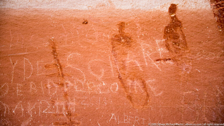 A broad filled figure, a narrow linear figure, and a dark branching motif appear on sandstone.