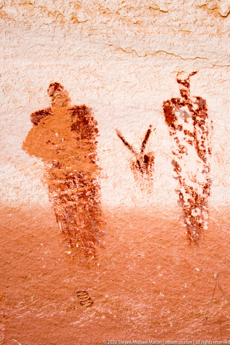 A tall outlined human-like figure and a smaller dark motif appear on pale sandstone.