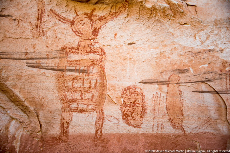 A row of faded painted figures follows a pale sandstone wall beneath a projecting ledge.