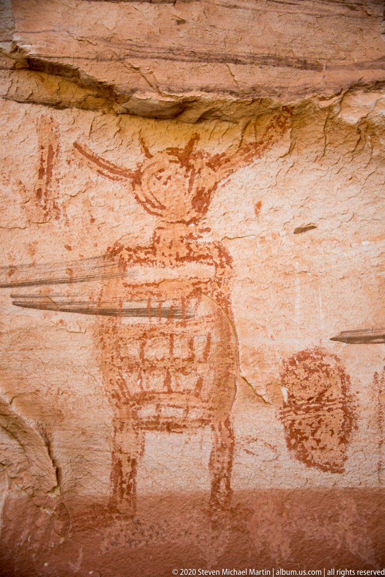 A dark mottled painted shape stands beside barely visible vertical figures on pale sandstone.