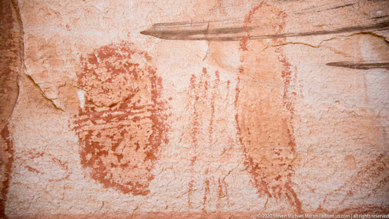 Faint elongated figures and geometric marks cross a shadowed sandstone wall above a dark ledge.