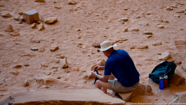 Thin horizontal sandstone layers form stepped ledges among desert shrubs beneath a blue sky.