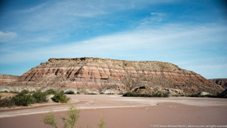 A flat-topped, red-and-cream-banded ridge rises beyond a pale basin and sparse shrubs.