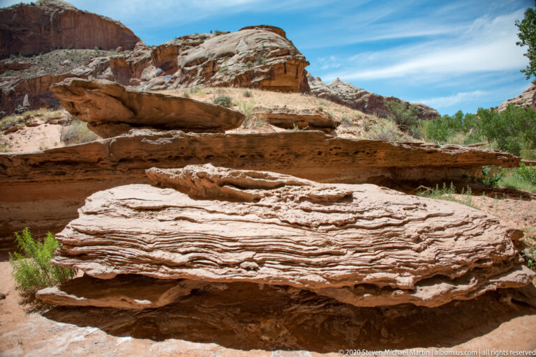 Thin horizontal sandstone layers form stepped ledges among desert shrubs beneath a blue sky.