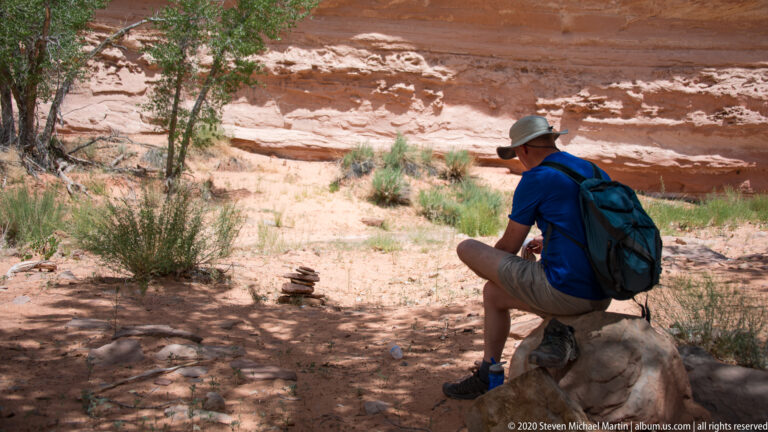 A seated hiker with a backpack rests beside a small stone stack beneath a sandstone wall.