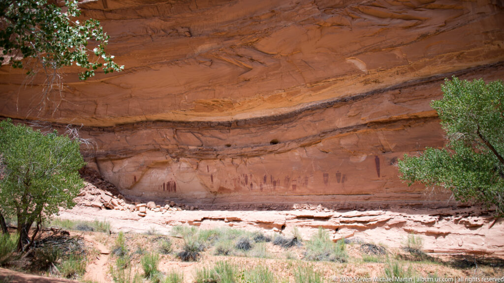 Dark painted figures line the lower sandstone wall beneath a broad ledge framed by shrubs.