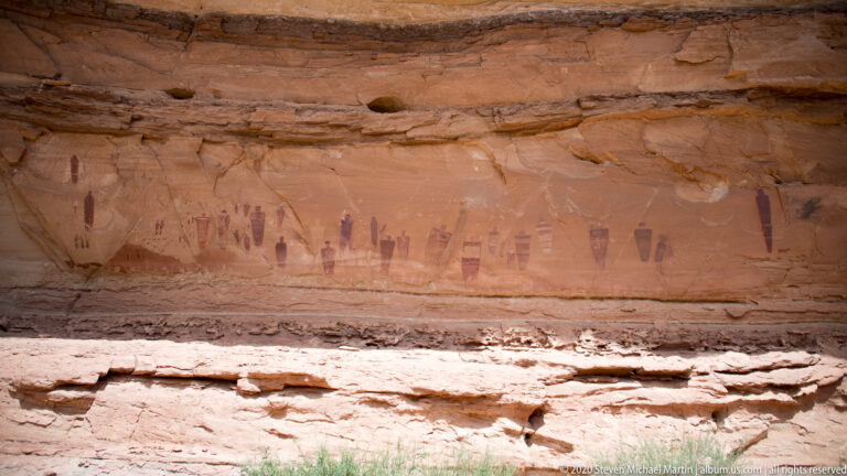 A row of dark upright and patterned forms spans a recessed sandstone panel above a pale ledge.