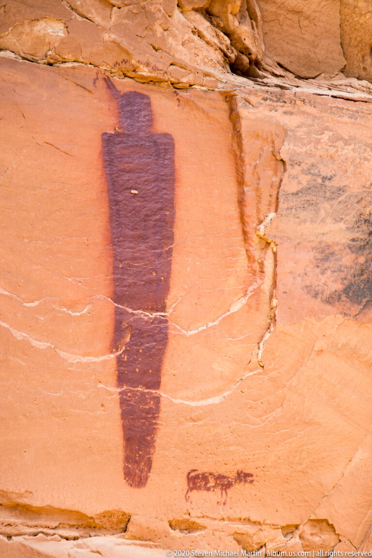 A tall tapered dark figure stands above a small horizontal painted motif on orange sandstone.