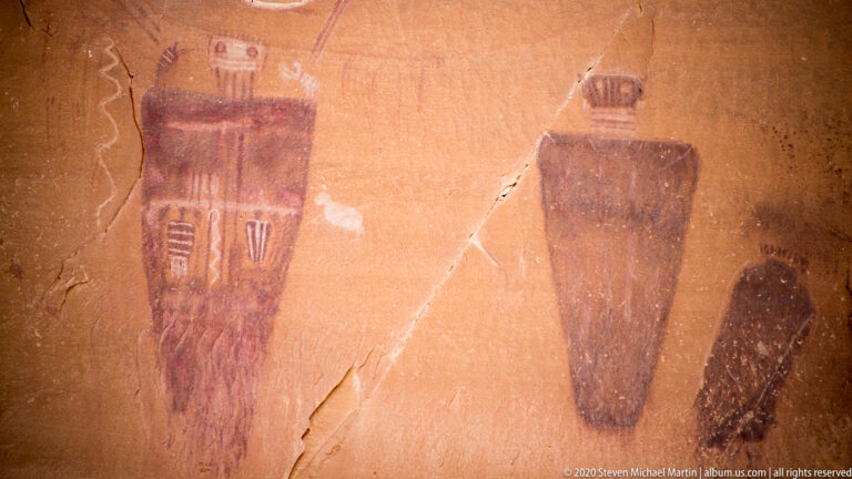 Three elongated painted figures stand on sandstone, including a patterned left form and a small figure at right.