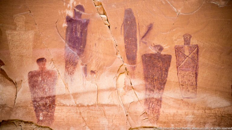 A group of tall painted figures crosses cracked sandstone, including a crosshatched form at right.