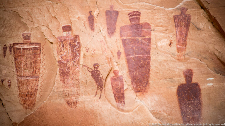 Large tapered figures and smaller forms share a weathered sandstone panel crossed by a pale line.