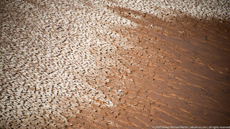 Pale cracked clay plates meet a diagonal strip of smaller fragments and dark sediment seams.