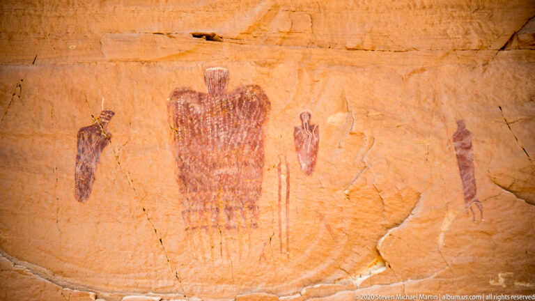 A large patterned figure stands between smaller upright forms on a cracked sandstone panel.