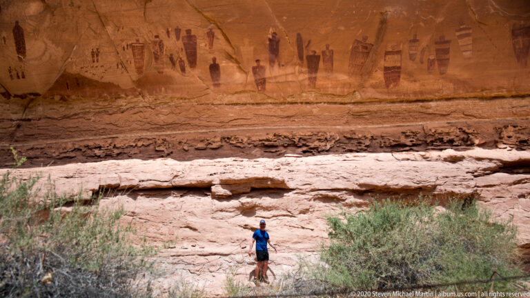 A small hiker stands below a long row of painted figures on a high sandstone wall.