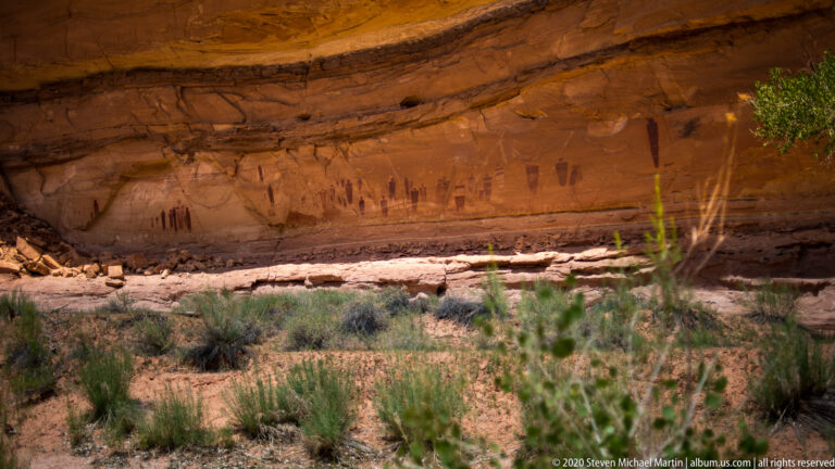 Dark pictographs line a shaded sandstone wall framed by trees and shrubs above sandy ground.