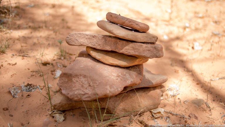 Flat reddish stones are stacked into a small cairn on a larger rock amid sandy ground and grass.