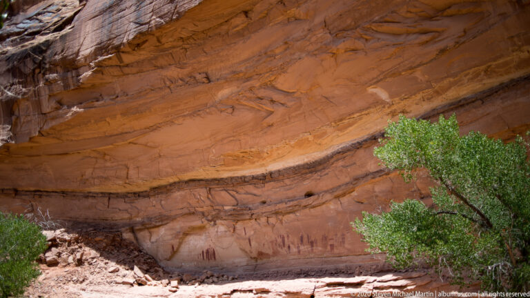 Dark figures line a shallow sandstone recess beneath layered rock, partly screened by a leafy tree.