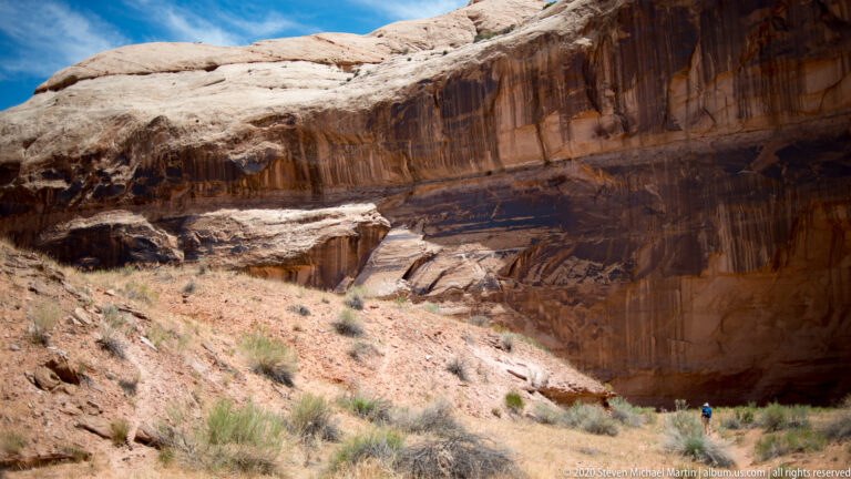 A small hiker stands below a high sandstone wall marked by dark streaks and painted forms.