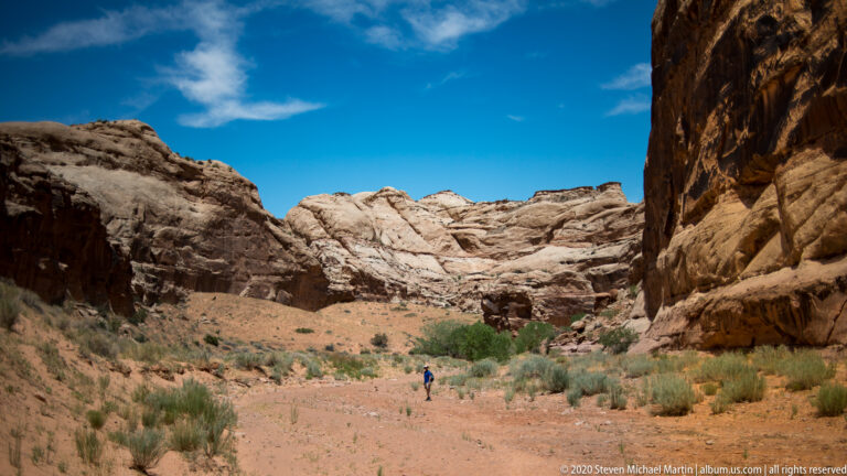 A visitor walks along a sandy canyon floor between pale rock formations and dark overhanging walls.