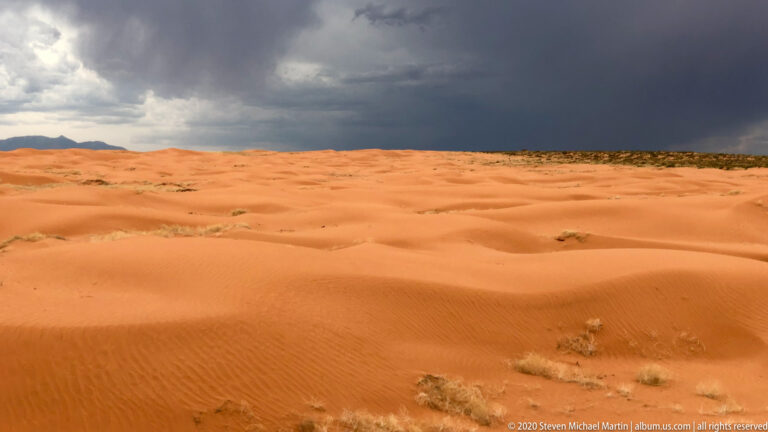 Orange dunes with sparse plants extend toward distant low mountains beneath a dark stormy sky.