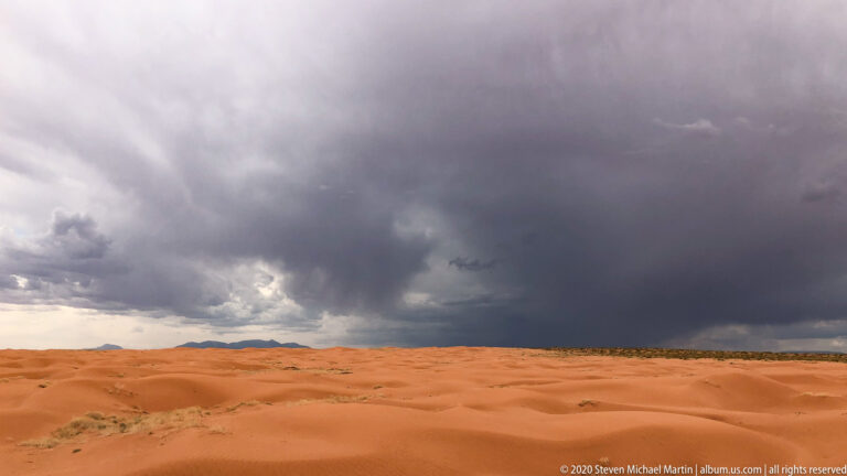Orange dunes and sparse shrubs lie beneath a broad gray storm cloud, with distant mesas on the horizon.