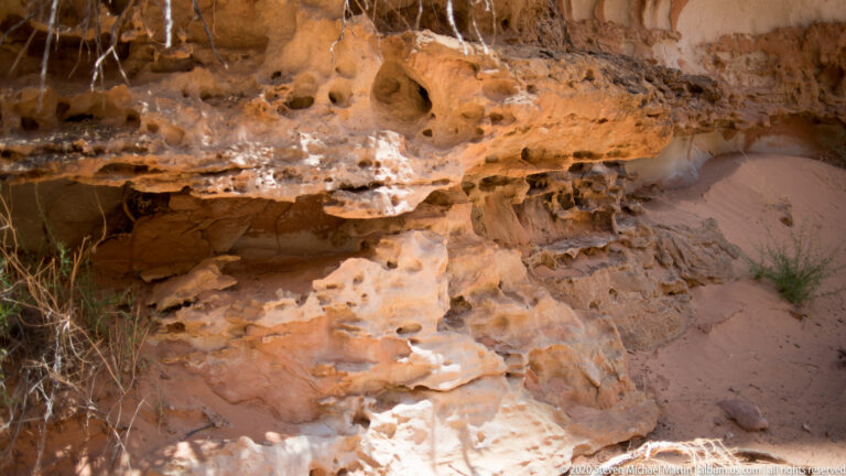 Pitted orange sandstone projects over a shaded sandy recess with sparse grasses below.