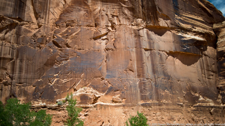 Long dark streaks mark a tall rust-colored sandstone cliff above green shrubs.