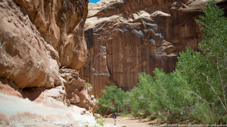 A lone hiker in blue stands on a sandy path between towering sandstone walls and green trees.