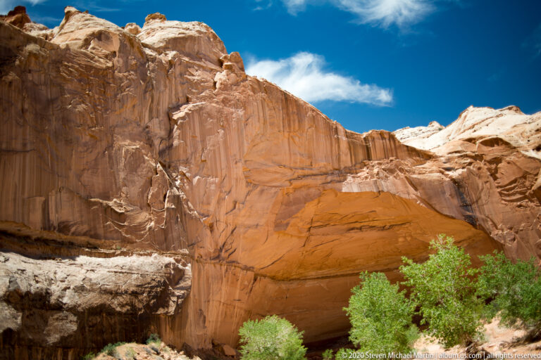 A broad orange sandstone alcove curves beneath pale fractured rock and blue sky, with shrubs at its base.